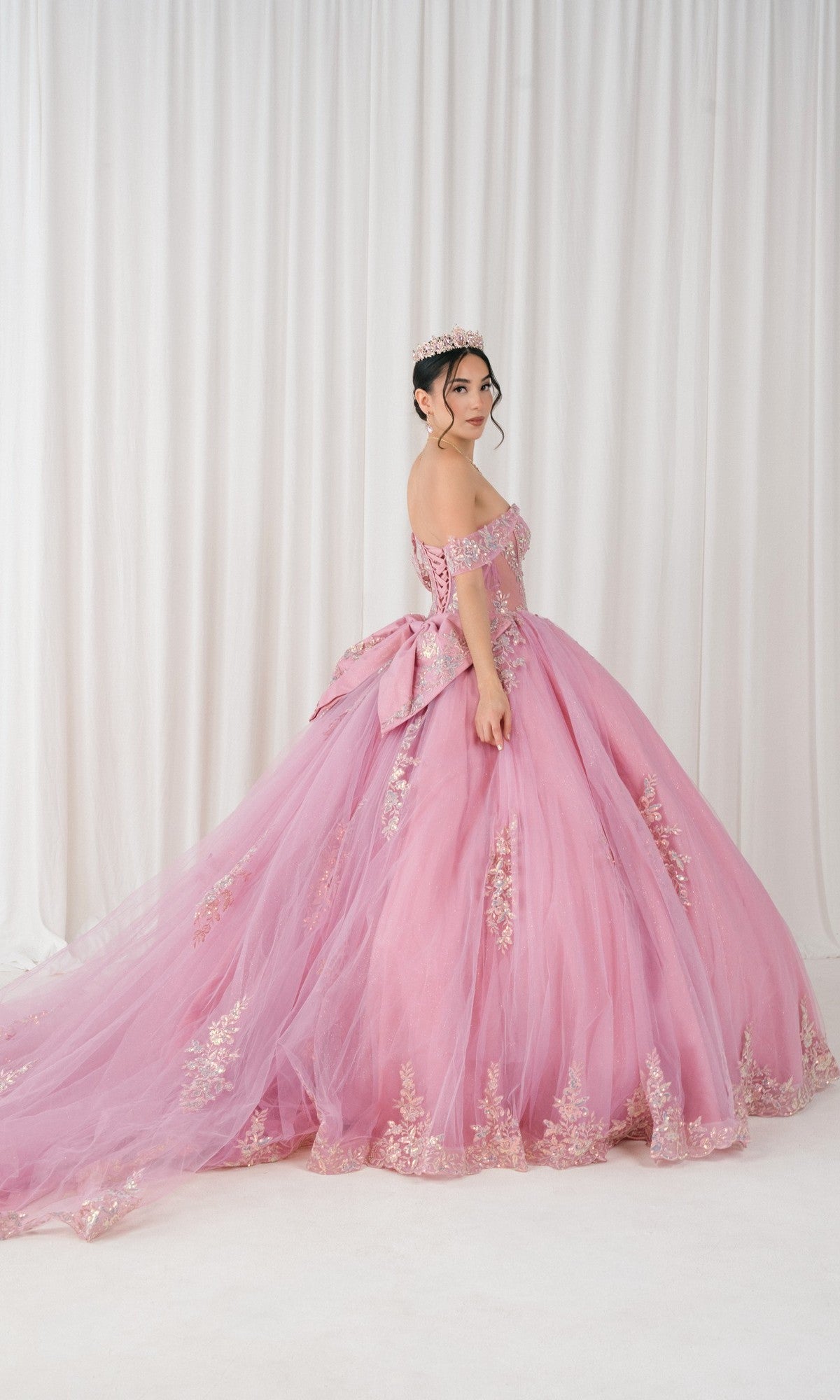 A young woman wearing the Quinceanera Dress 8405JQ By Cinderella Couture with gold detailing and a matching tiara stands in front of white curtains, looking over her shoulder.